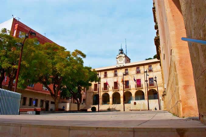 Vista de la fachada del Ayuntamiento de Dénia con su torre del reloj y arcos, rodeada de árboles en la Plaza de la Constitución