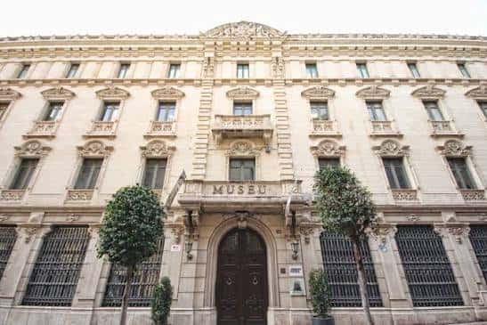 Entrada frontal del museo con letrero 'MUSEU' y árboles decorativos a los lados