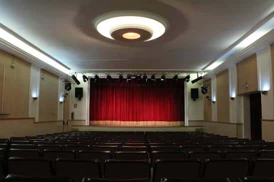 Interior del teatro con butacas negras y cortina roja en el escenario