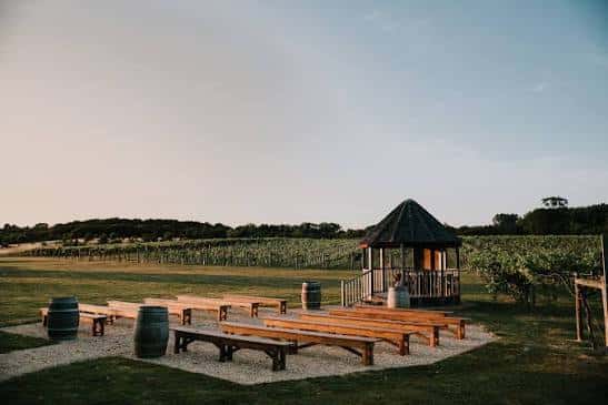 Área de celebración en viñedo con bancos de madera y gazebo bajo el cielo al atardecer