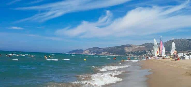 Playa de Castelldefels con personas disfrutando del agua y veleros en la orilla
