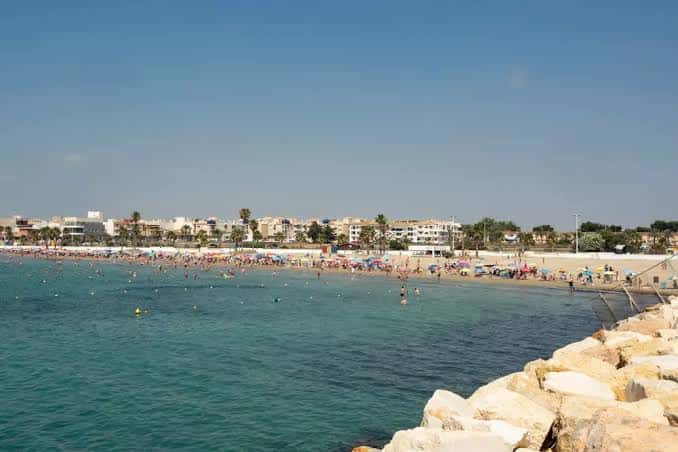 Playa concurrida con bañistas, sombrillas y rompeolas de piedra en primer plano