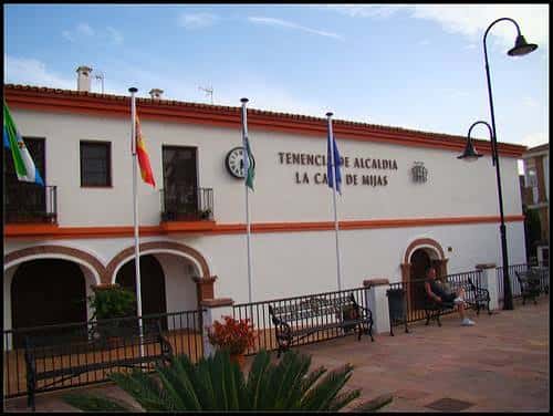 Fachada frontal del edificio con banderas ondeando y bancos en la plaza