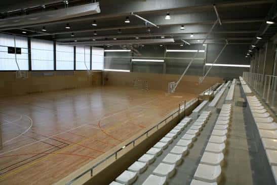 Interior del pabellón deportivo con pista de madera y gradas blancas