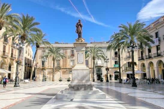 Plaza central con estatua en el centro, rodeada de palmeras y edificios históricos con arcos