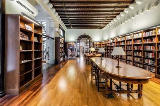 Amplio salón de lectura con mesa central, lámparas y estanterías llenas de libros