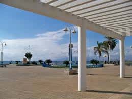 Perspectiva bajo el pórtico blanco de la plaza, mostrando el paseo marítimo y el mar en el horizonte