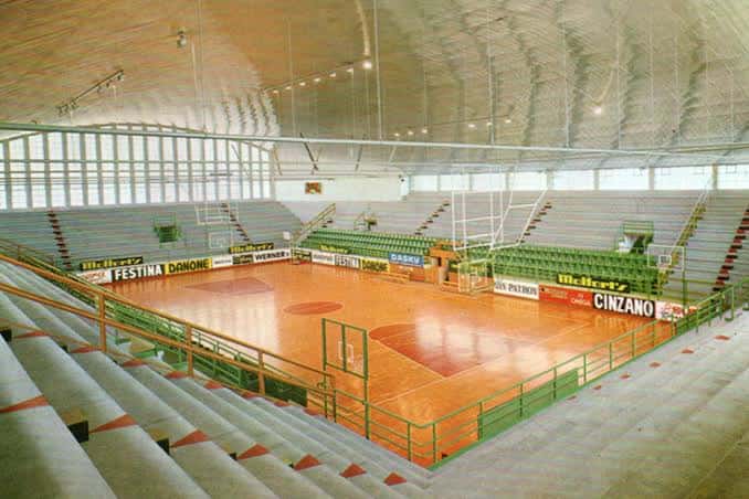 Interior del pabellón con gradas verdes y pista de madera, vista desde las alturas