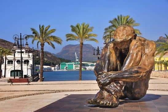 Vista completa de la escultura 'El Zulo' frente al mar con palmeras y montañas al fondo