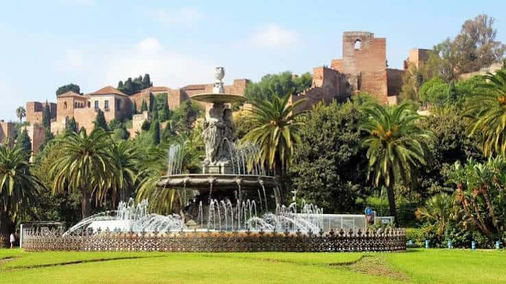 Fuente ornamental en primer plano con la Alcazaba al fondo entre palmeras y jardines