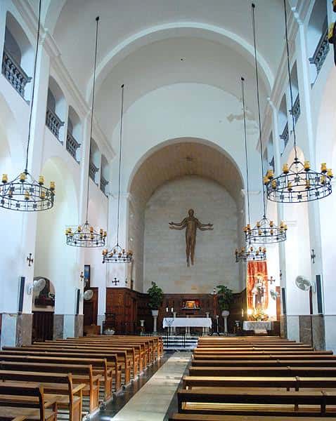 Interior de la nave con bancos de madera, candelabros colgantes y crucifijo en el altar