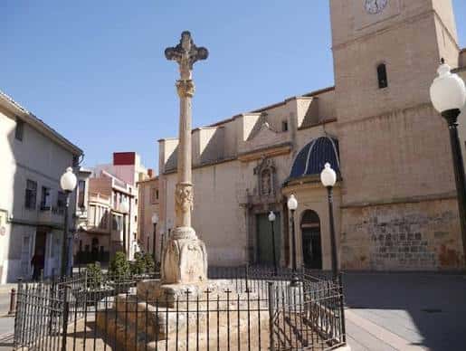Detalle de la cruz de piedra en el centro de la plaza con la fachada lateral de la iglesia al fondo