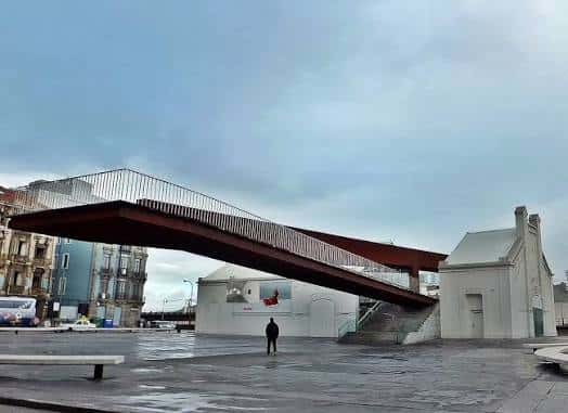 La pasarela de La Grapa sobre la plaza, con el edificio blanco y el cielo nublado de fondo