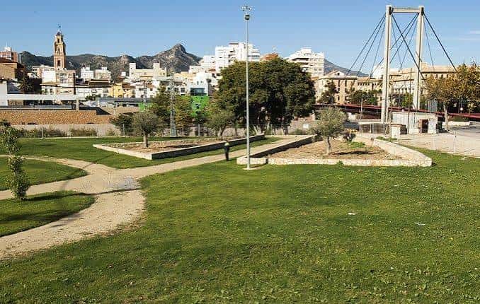 Áreas verdes y senderos del parque con el puente colgante y la ciudad de Gandia al fondo