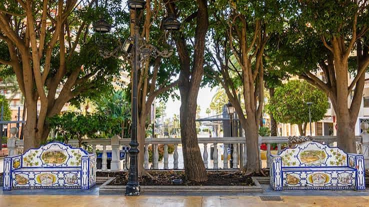 Dos bancos de cerámica bajo árboles frondosos en la zona peatonal del parque