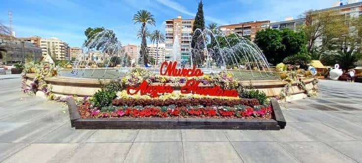 Plaza Circular decorada con flores y letrero festivo 'Murcia Alegre y Humana' frente a la fuente