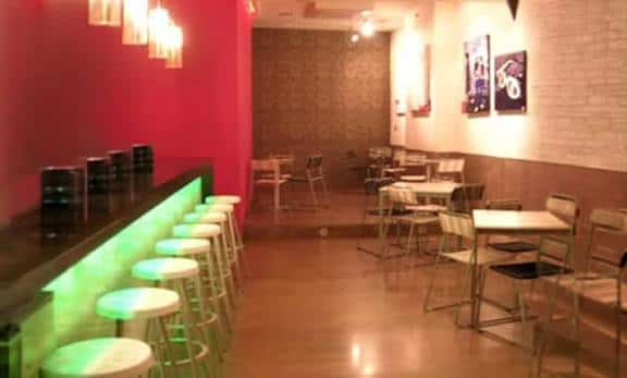 Vista del bar con barra iluminada en verde y mesas con sillas metálicas