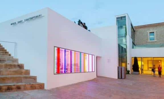Fachada del MACE al atardecer con ventana iluminada en colores vibrantes y siluetas en la terraza