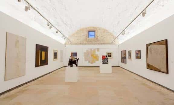 Interior de sala de exposiciones con bóveda de piedra, obras colgadas y suelo de piedra clara