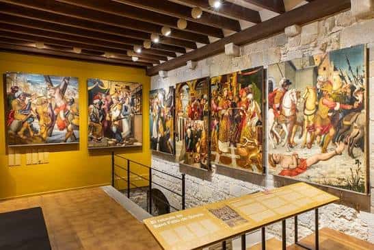 Exposición de pinturas históricas en una sala con paredes amarillas y vigas de madera