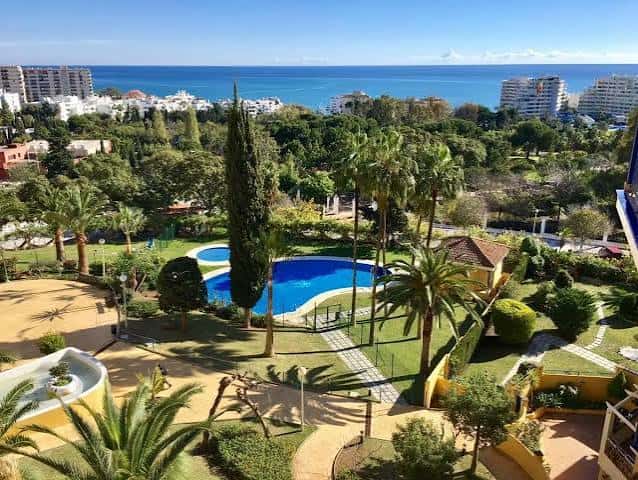Vista elevada del parque con piscina azul, palmeras y el mar Mediterráneo al fondo
