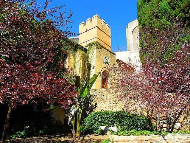 Vista de la torre y muros de los Jardines de la Galera rodeados de árboles de flores rojas