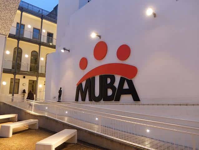 Fachada moderna del MUBA con logotipo rojo y negro iluminado al atardecer