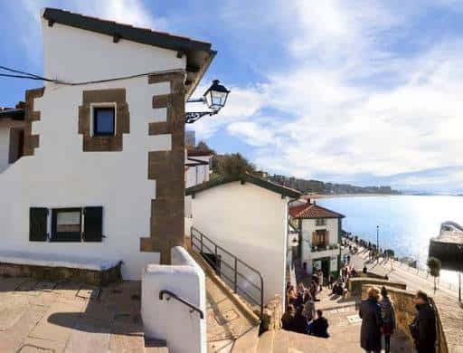 Escalinata con gente caminando hacia el mar, junto a una casa blanca con detalles en piedra