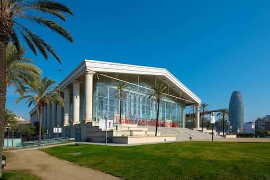Vista exterior del Teatre Nacional de Catalunya con fachada de cristal y columnas, bajo un cielo azul despejado