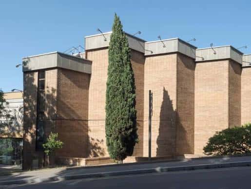 Vista lateral del edificio con fachada de ladrillo y ciprés alto en primer plano