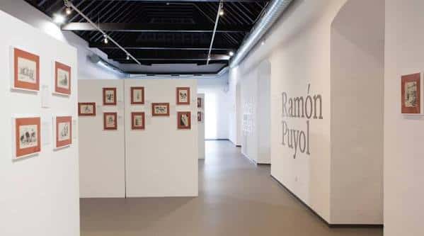 Espacio expositivo con paneles blancos, obras enmarcadas y nombre del artista en la pared