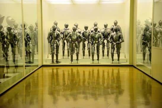 Exposición de armaduras medievales en el Museo del Ejército, con múltiples piezas exhibidas en vitrinas iluminadas y reflejos en el suelo pulido