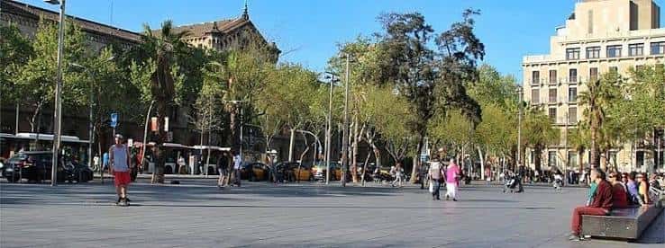 Plaza de la Universidad en Barcelona con árboles, personas caminando y edificios históricos al fondo