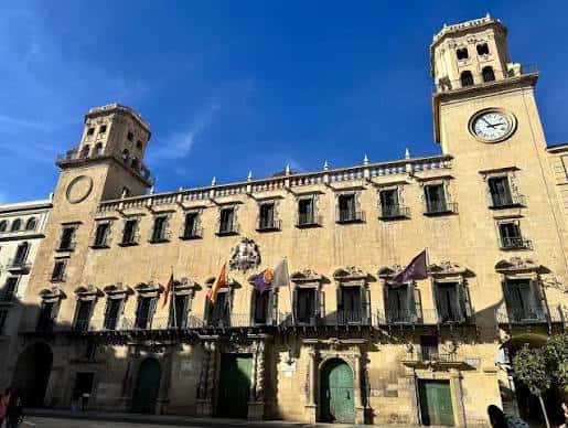 Fachada principal del Ayuntamiento de Alicante con torres gemelas y banderas bajo cielo azul