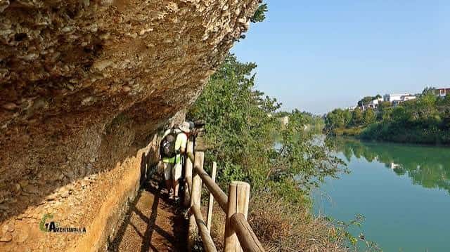 Senderistas caminando por un sendero junto al río con un acantilado rocoso