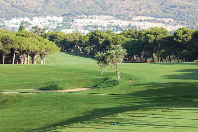 Amplia extensión del campo de golf con árboles dispersos y vista de la ciudad en la ladera de la montaña