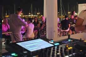 Escena de evento con DJ y asistentes disfrutando en la terraza del Moll de Costa