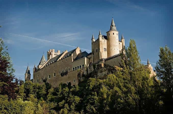 El Alcázar de Segovia en primer plano rodeado de árboles verdes bajo un cielo despejado