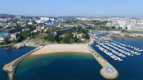 Vista aérea panorámica de la Playa de Oza mostrando su forma de media luna, el puerto deportivo y la ciudad de A Coruña al fondo