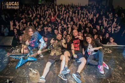 Banda posando en el escenario con público de fondo haciendo señas de rock