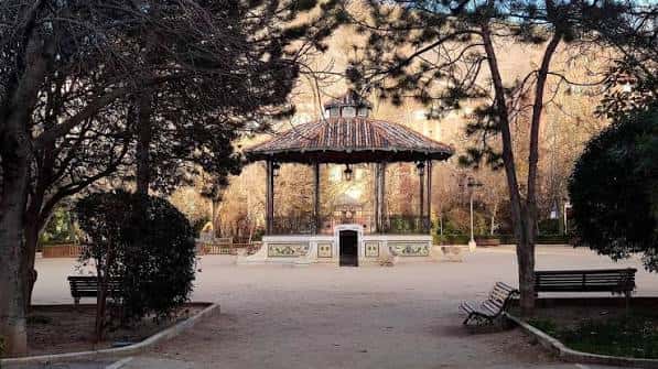 Kiosco del parque enmarcado por árboles en un día soleado, con bancos vacíos en primer plano