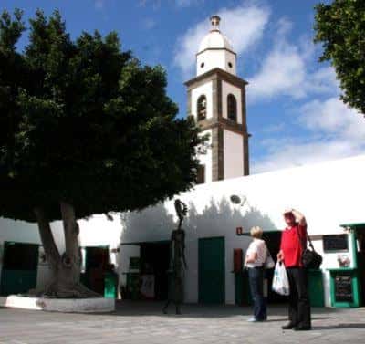 Plaza exterior de La Recova con árboles y la torre de la iglesia al fondo