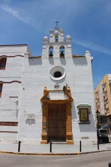Fachada exterior de la Capilla de la Caridad en Algeciras, con su característica torre campanario y puerta de madera bajo un cielo azul