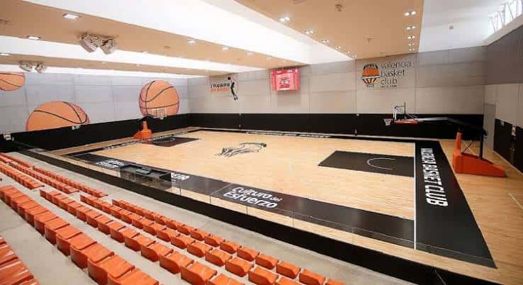 Pista de baloncesto interior con gradas naranjas, logotipos del club y diseño deportivo profesional