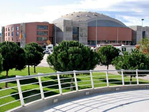 Vista frontal del recinto deportivo desde un puente peatonal con árboles en primer plano