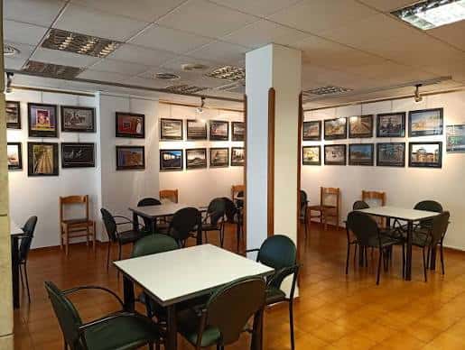 Sala de exposiciones con mesas, sillas y cuadros enmarcados en las paredes