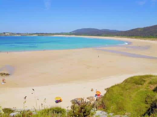 Playa amplia de arena dorada con aguas cristalinas y dunas verdes en primer plano