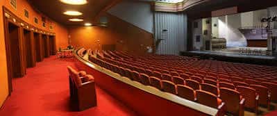 Interior del teatro con butacas rojas, pasillo central y escenario iluminado