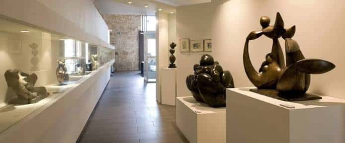 Galería de exposiciones con esculturas de bronce sobre pedestales blancos y vitrinas laterales