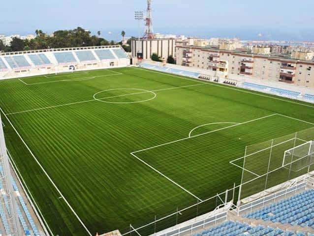 Área central del estadio mostrando el césped y las gradas azules con el mar al fondo